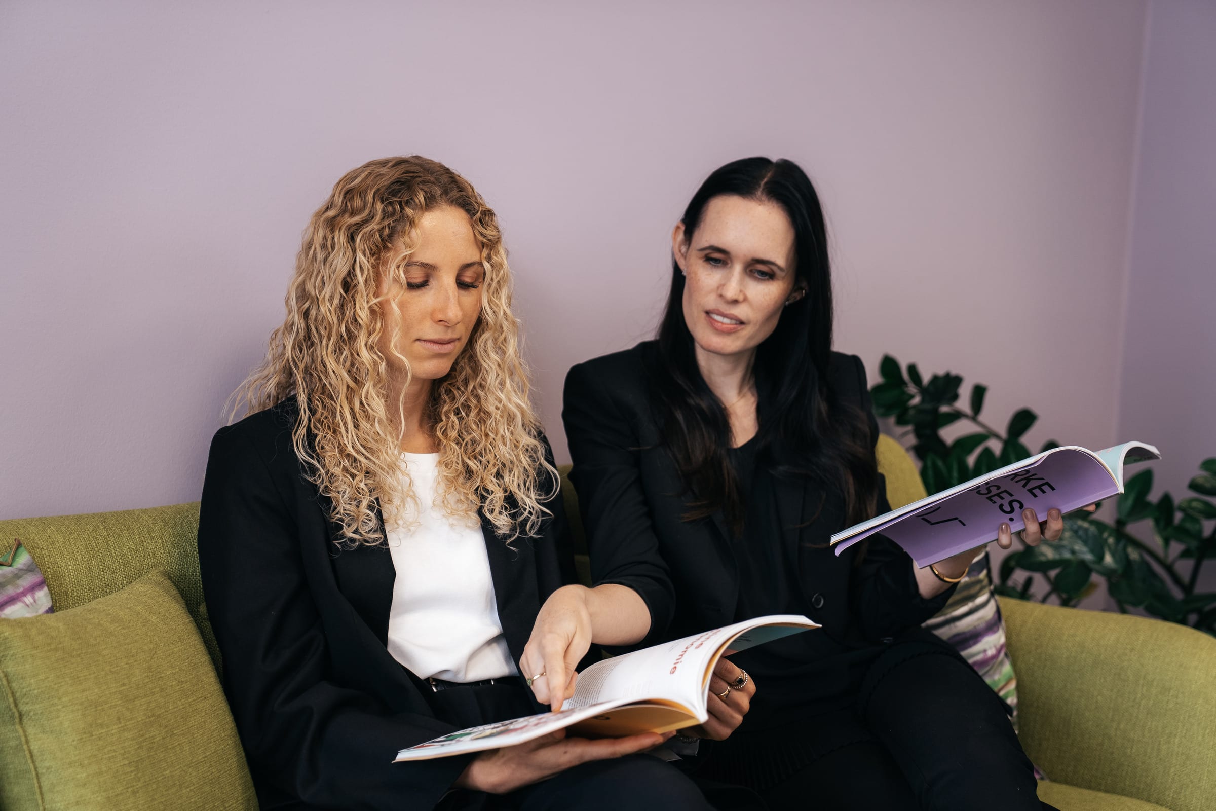 Anthea Wagner und Jessica Ryan-Tietz sitzen auf einem grünen Sofa und lesen gemeinsam in einer Zeitschrift