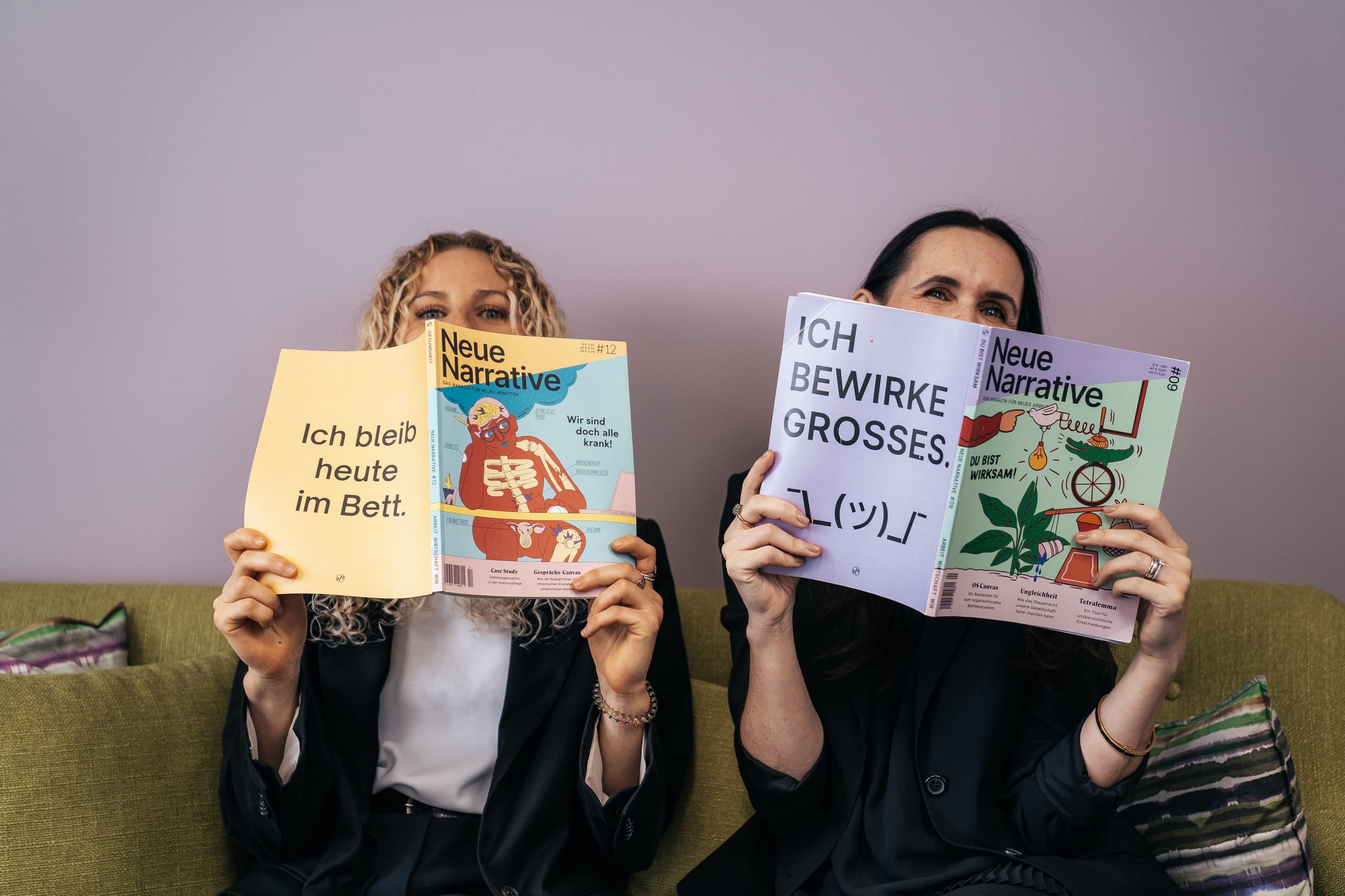 Anthea Wagner und Jessica Ryan-Tietz halten sich Zeitschriften vors Gesicht, auf den Titeln steht „Ich bleib heute im Bett“ und „Ich bewirke Großes“