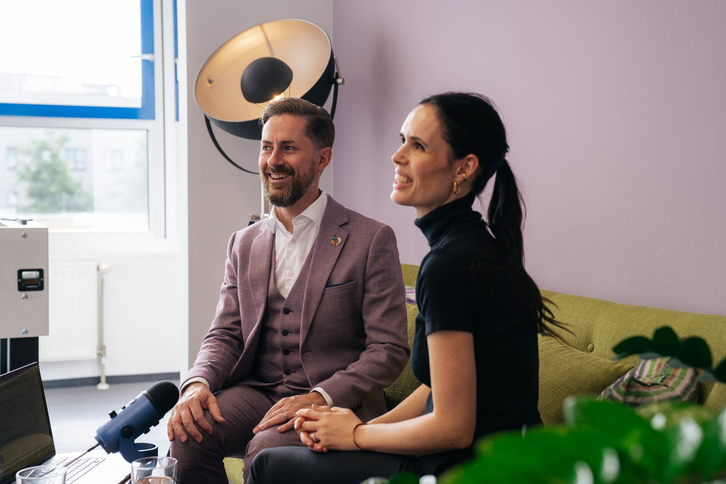 Rainer Karcher und Jessica Ryan-Tietz sitzen lachend nebeneinander auf einem grünen Sofa, vorne ein Mikrofon