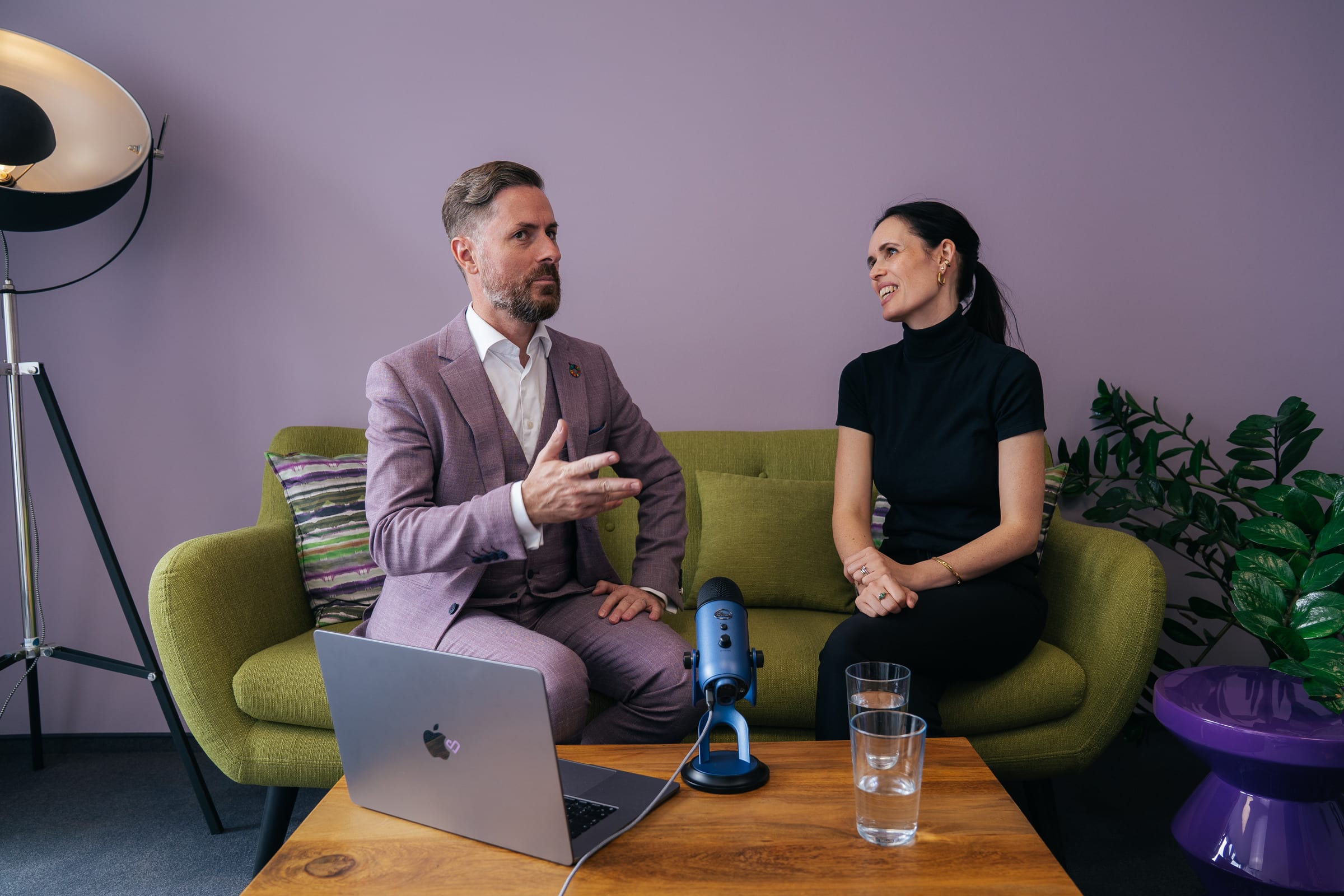 Rainer Karcher erklärt etwas mit erhobener Hand, Jessica Ryan-Tietz hört ihm lächelnd zu; beide sitzen auf einem grünen Sofa, vorne Laptop und Mikrofon