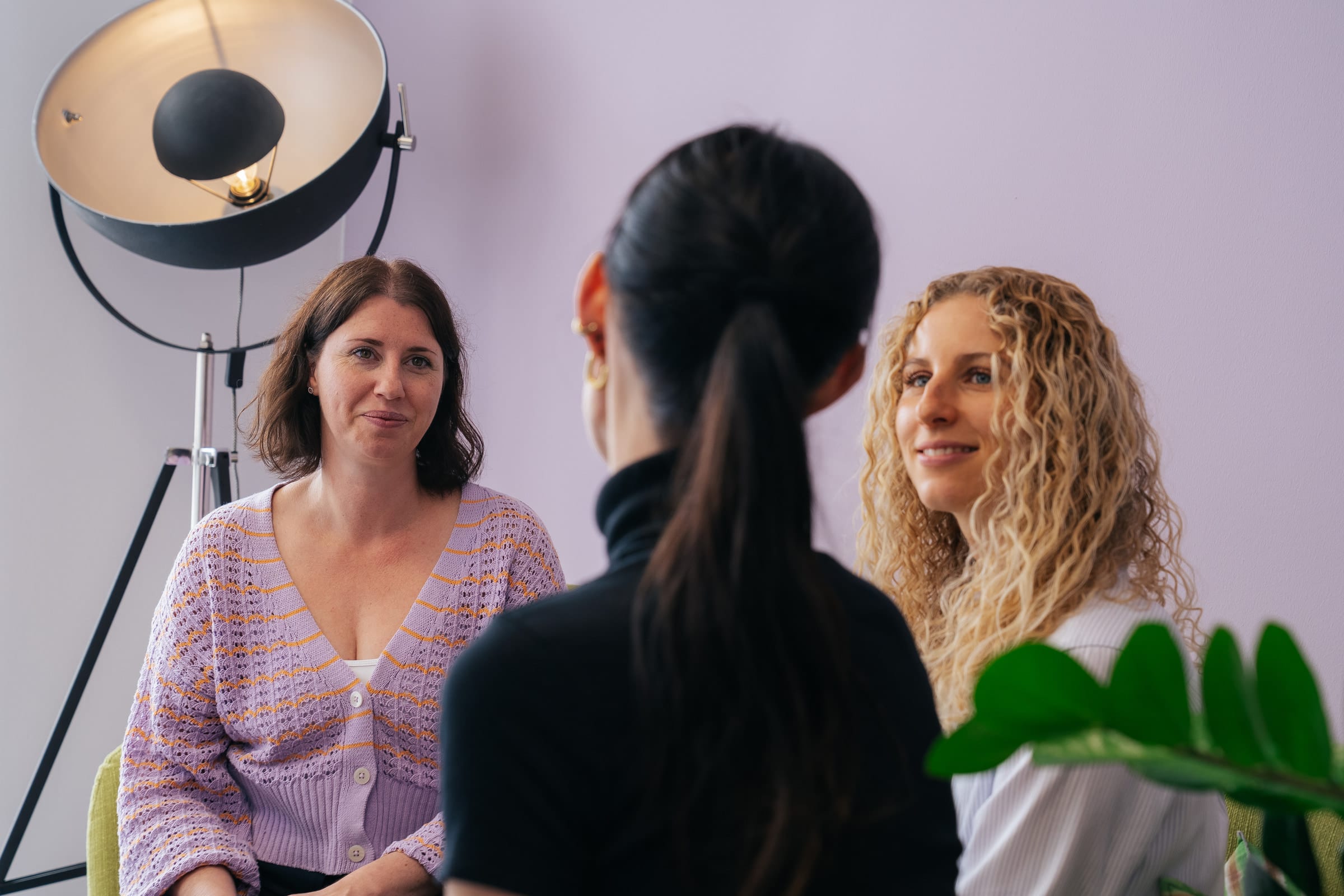Stefanie Karcher und Anthea Wagner sitzen vor einer lila Wand und hören Jessica Ryan-Tietz zu, die mit dem Rücken zur Kamera sitzt