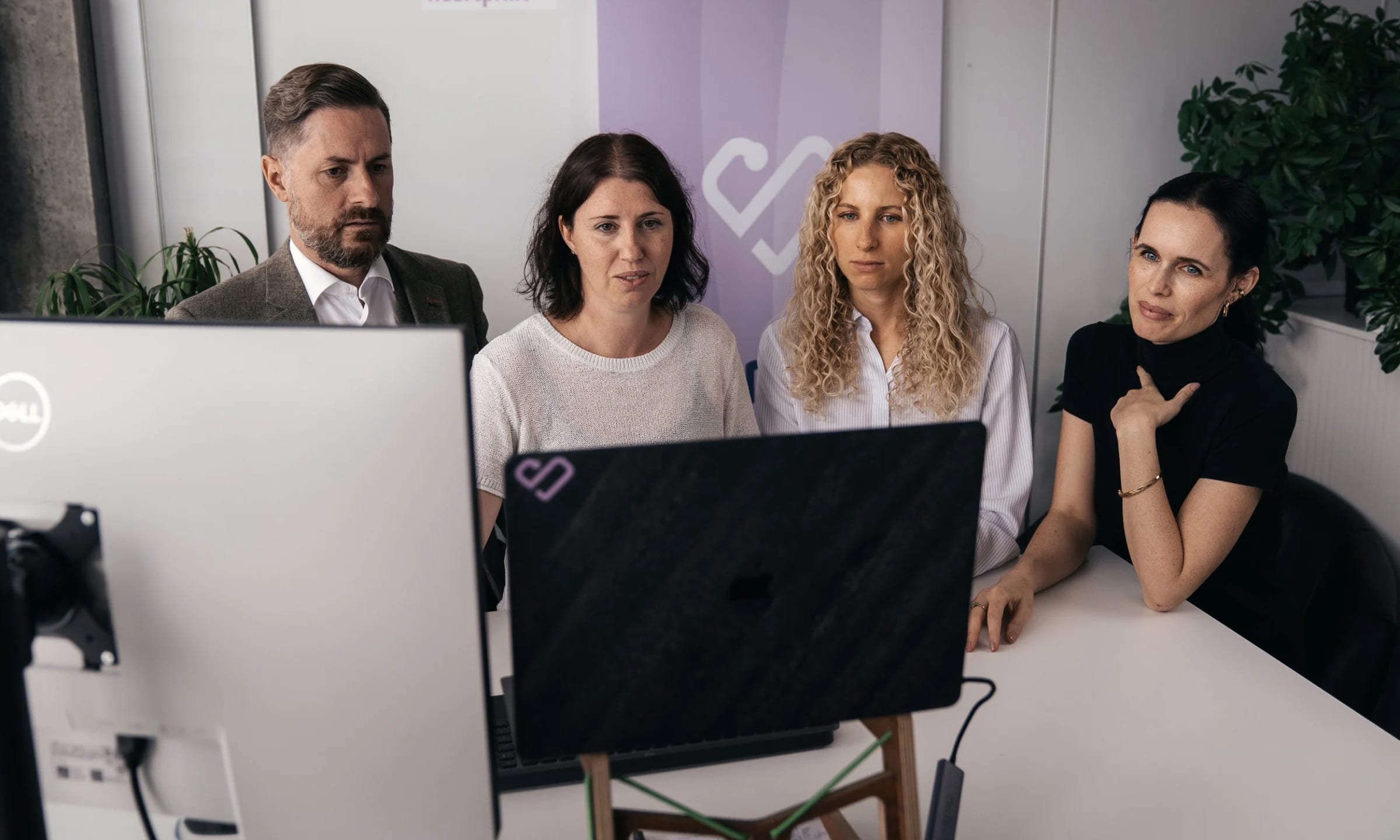 Rainer Karcher, Stefanie Karcher, Anthea Wagner und Jessica Ryan-Tietz schauen gemeinsam auf einen Laptop, hinter ihnen ein Roll-up mit dem Heartprint-Herz