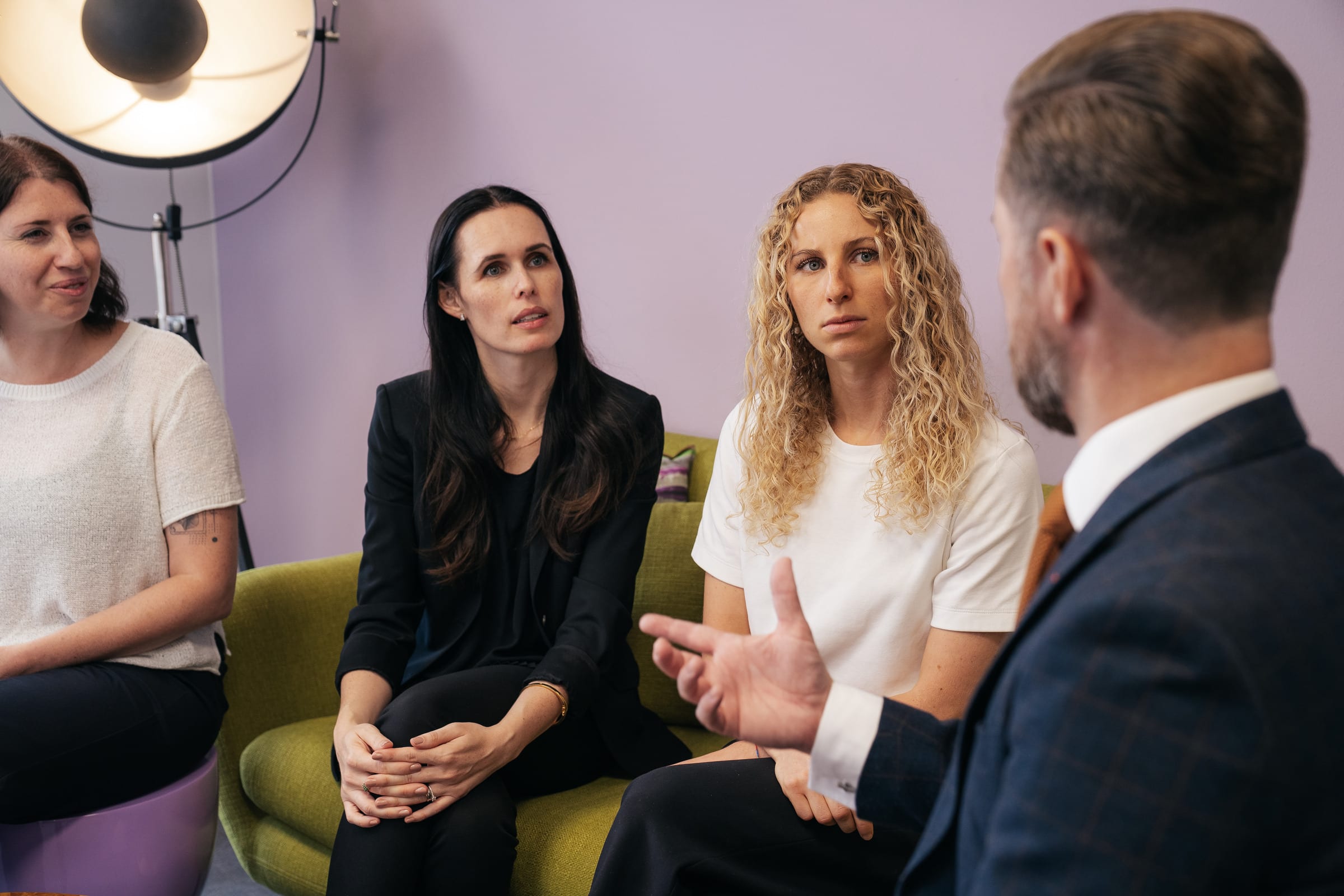 Rainer Karcher, von hinten zu sehen, erklärt mit offener Hand; Stefanie Karcher, Jessica Ryan-Tietz und Anthea Wagner hören ihm auf einem grünen Sofa vor lila Wand zu