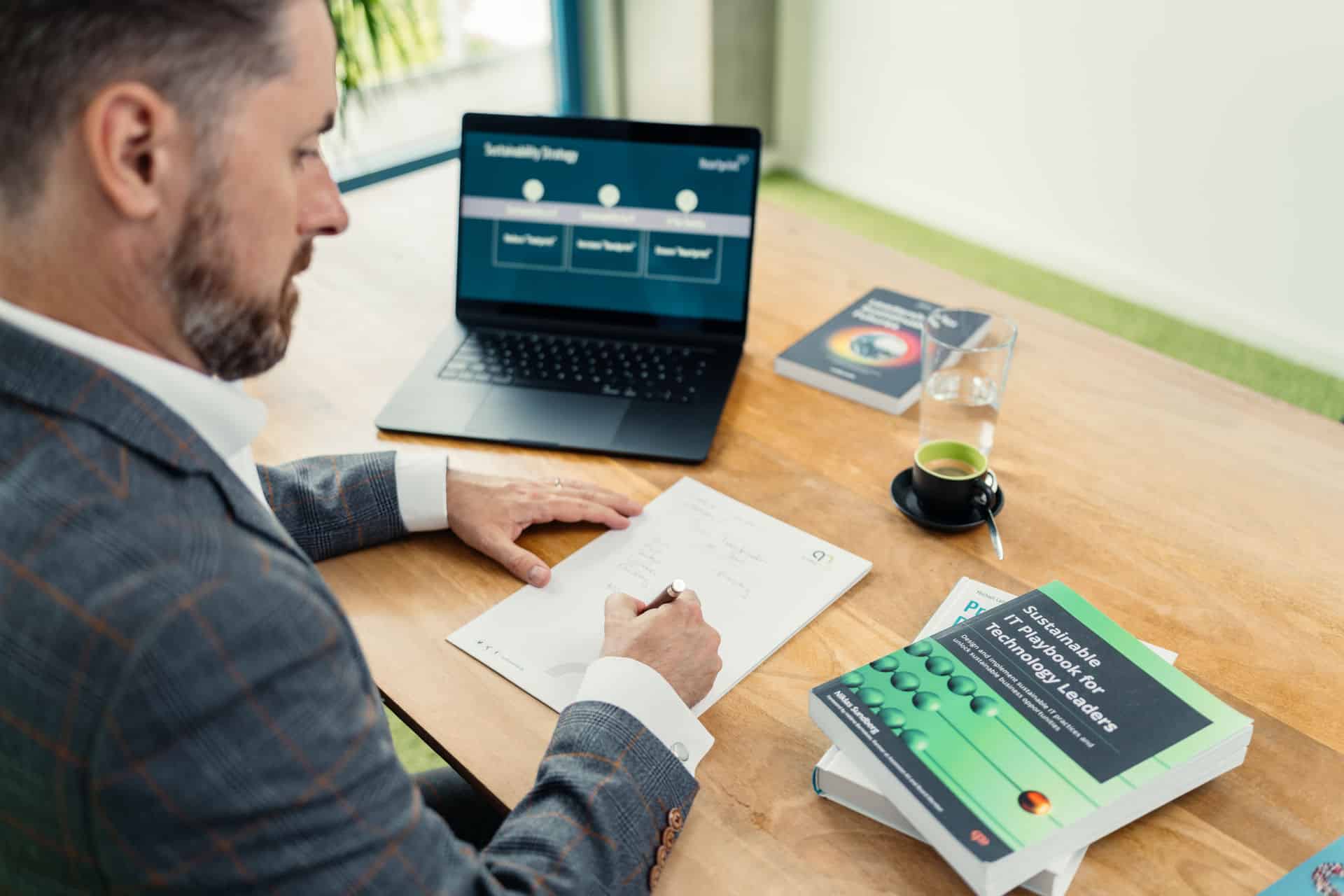 Rainer Karcher sitzt am Schreibtisch und schreibt etwas. Dabei steht der Laptop offen und es liegen Bücher zu Sustainability auf dem Tisch.